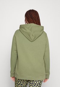 Vihreä oliivinvärinen huppari, joka on valmistettu puuvillasta. Siinä on suuri huppu ja ribbireunat, yhdistettynä kukkakuosisiiin housuihin.