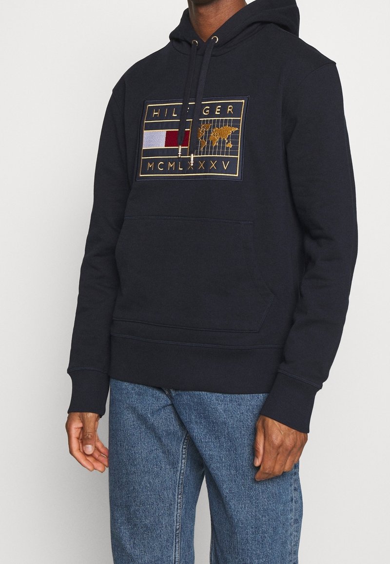 Námořnicky modrá mikina s kapucí, přední kapsou, stahovací šňůrkou a vyšívaným logem ve zlaté, červené a bílé barvě s motivem mapy světa.