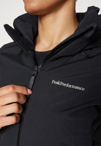 Schwarze Softshell-Jacke mit hohem Kragen, Reißverschluss vorne und gesticktem "PeakPerformance"-Logo auf der Brust. Verfügt über verstellbare Manschetten.