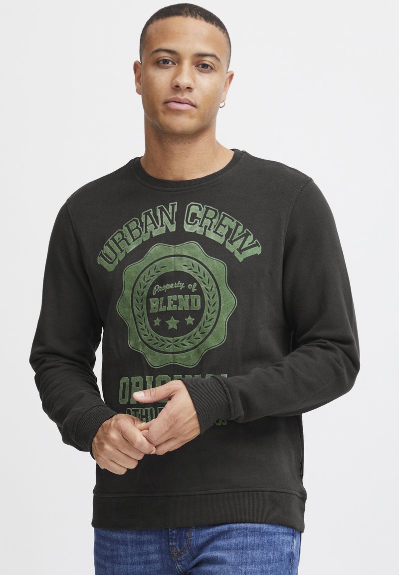 Sudadera negra de cuello redondo con texto gráfico y emblema verde, mangas largas, tela texturizada. Combinada con jeans de mezclilla azules.