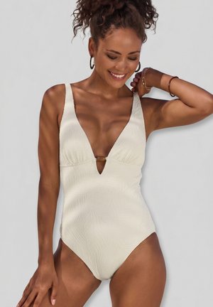 Smiling woman with curly hair wears textured white one-piece swimsuit with deep V-neck and ring detail, posing against plain light background.