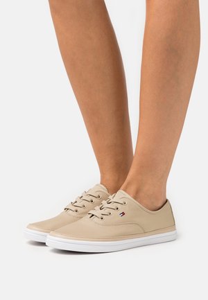Zapatillas bajas de color beige con suelas y cordones blancos, usadas en pies desnudos sobre un fondo claro y uniforme.