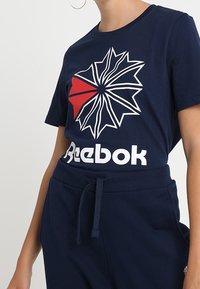 Námořnická modrá bavlněná tričko s velkým grafickým prvkem s červeným trojúhelníkem a bílým logem "Reebok" na dolním okraji. Krátké rukávy, volný střih.