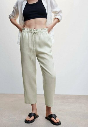 Mujer con pantalones beige con cordón, top negro corto, camisa blanca abierta y sandalias negras, de pie con las manos en los bolsillos.