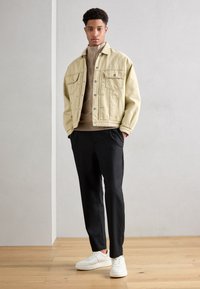 En ljusblå denimjacka med knäppte fickor, beige tröja, svarta byxor och vita sneakers, står mot en neutral bakgrund.