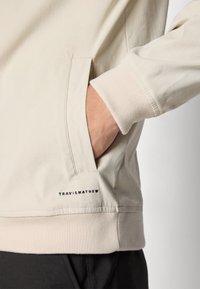 Cream-colored sweatshirt with a relaxed fit, featuring a side pocket and ribbed cuffs. The brand name "TRAVISMATHEW" is displayed near the hem.