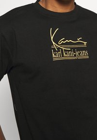Camiseta de algodón negra con un diseño dorado que incluye el texto "Kani" y "karl kani-jeans". Cuello redondo y mangas cortas, textura suave.