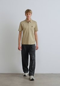 Bézs színű galléros póló, bal oldalon melltartóval és logóval, sötétszürke nadrággal és fehér tornacipővel párosítva. Sima anyag textúra.