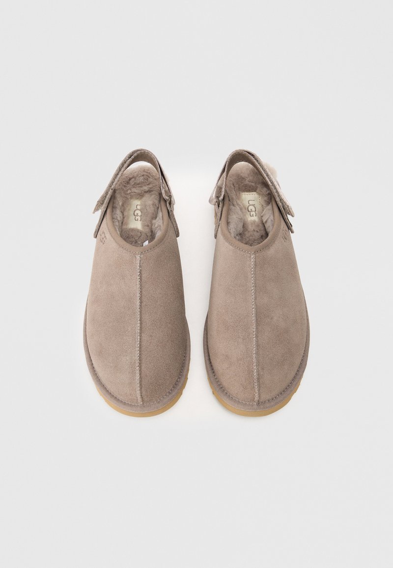 Par de zapatillas de ante beige tipo slip-on con forro de piel de pelo, correas ajustables y plantillas de marca sobre un fondo claro.