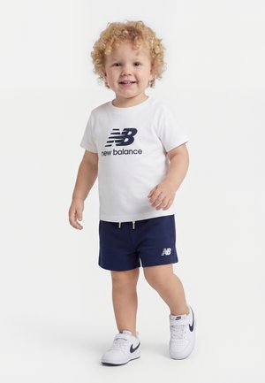 Bambino con capelli biondi ricci che indossa una T-shirt bianca New Balance, pantaloncini blu navy e sneakers bianche Nike, sorridente e che cammina in avanti.
