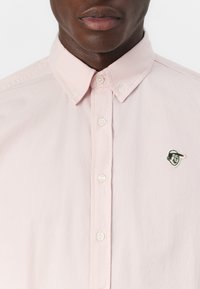 Light pink button-up shirt with a pointed collar, featuring white buttons and a black and white embroidered logo on the left chest.