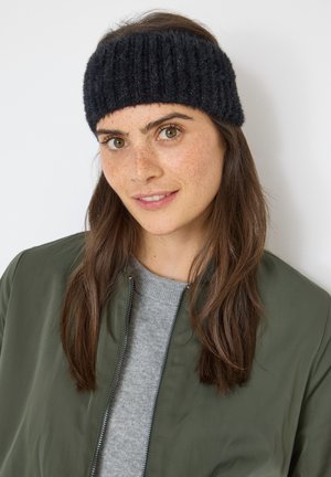 Bandeau tricoté noir à texture côtelée, porté avec une veste bomber vert olive et un pull gris, doté d'un détail zippé.