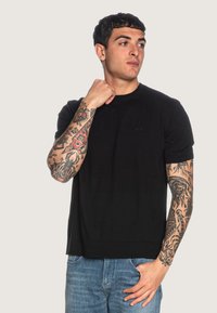 Maglietta nera a maniche corte a girocollo in cotone, con un logo discreto sul petto. La vestibilità è rilassata, abbinata a jeans blu.