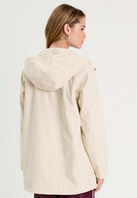 Veste imperméable beige avec une capuche, présentant une coupe droite, des manches longues et un matériel minimal. Texture lisse, design simple.