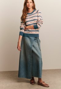 Vrouw in een gebreide trui met patroon, lange denim wikkelrok en bruine Mary Jane ballerina's, staand tegen een effen beige achtergrond.