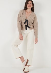 Beige cable-knit cardigan with a deep V-neck and black tie belt, paired with cream wide-leg pants and beige sneakers with pink accents.