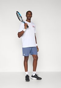 Hvid T-shirt med korte ærmer med rød og sort grafik, blå atletikshorts med hvidt Adidas-logo, sorte sneakers, holder en tennisketcher.