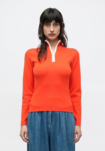 Pull-over en maille rouge avec une demi-fermeture éclair et des détails blancs, doté d'un corps ajusté et de manches longues, accompagné d'un pantalon en denim bleu ample.