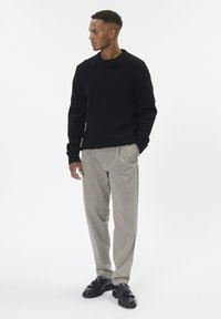 Vīrietis melnā džemperī, beidzos biksēs un melnās mokās, stāv ar rokām kabatās, raugoties pa kreisi uz vienkāršas fona.