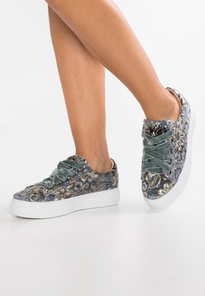 Jalat, joissa on sinivalkoiset samettiset tennarit, joissa on paksut valkoiset pohjat ja leveät vihreät sametinauhat valkoisella taustalla.