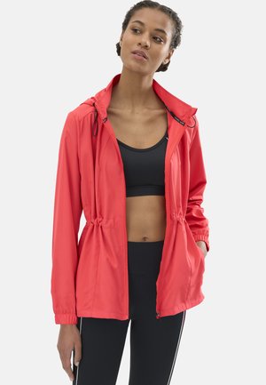 Mujer joven con chaqueta coral con cremallera sobre un sujetador deportivo negro y leggings negros con franjas blancas en los costados, de pie con una mano en el bolsillo.