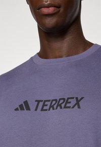 Fioletowa koszulka bawełniana z krótkim rękawem i okrągłym dekoltem. Posiada czarne logo "TERREX" i trzy czarne paski po lewej stronie.