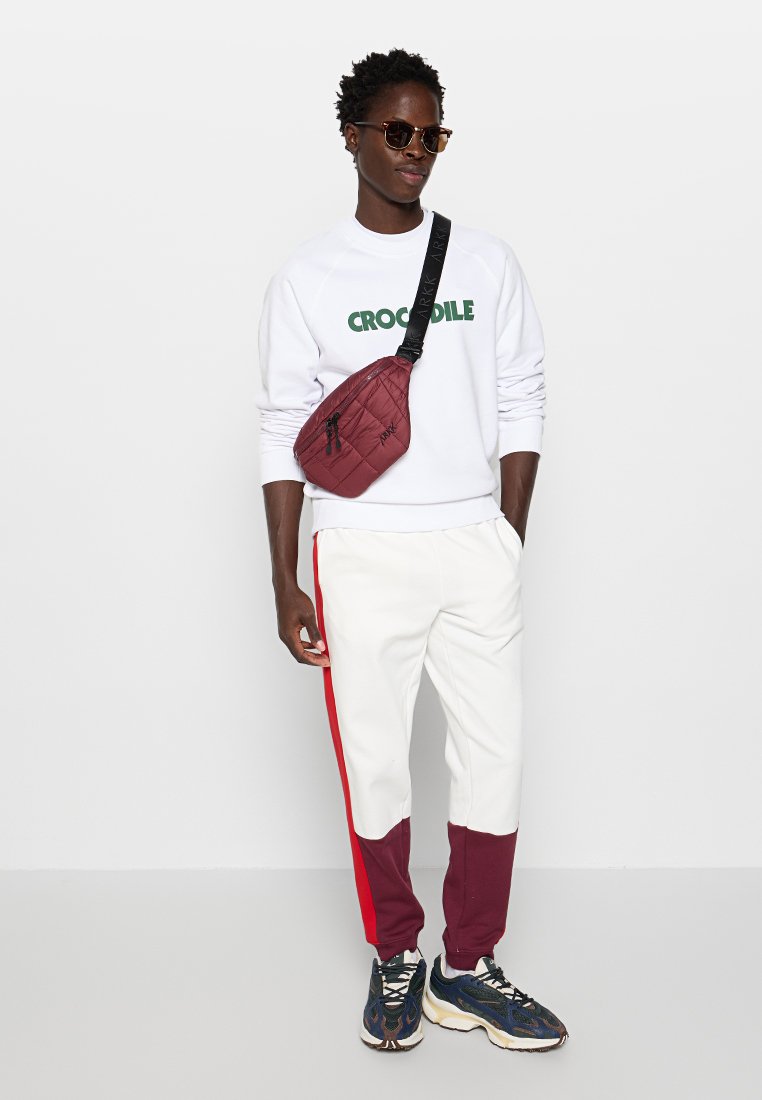 Weißes Sweatshirt mit grünem "CROCODILE"-Logo, weiße und granatrote Jogginghose mit roten Seitenstreifen, granatrote Umhängetasche und gemusterte Sneaker.