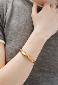 Brazalete de oro trenzado reposando sobre un brazo de piel clara, con un acabado suave y brillo reflectante, llevado sobre una camisa gris.