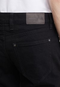 Jeans in denim nero con una texture liscia, dotati di una toppa in pelle grigia sul retro della vita e tasche a doppia cucitura.