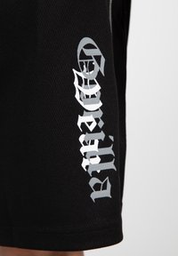 Zwarte mesh shorts met een gestructureerde afwerking. Voorzien van gedurfde zilveren letters aan de zijkant, wat een duidelijk contrast creëert. Eenvoudig ontwerp, knielang.