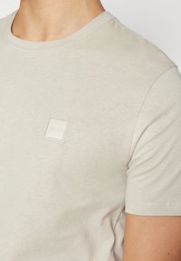 Béžové bavlnené tričko s okrúhlym výstrihom, krátkymi rukávmi a malou bielou látkovou nášivkou na ľavej strane hrudi.