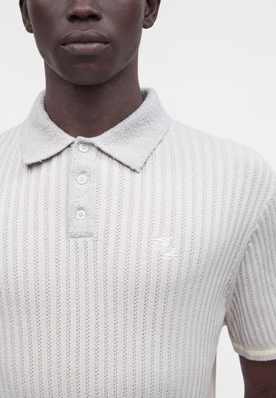 Personne à la peau foncée portant un polo en maille blanche avec un col texturé et des rayures verticales en maille ajourée, boutons sur le devant.