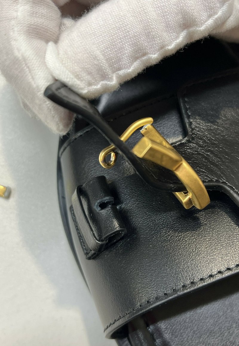 Main gantée tenant une sangle en cuir noir avec une boucle métallique dorée cassée sur un objet en cuir noir.