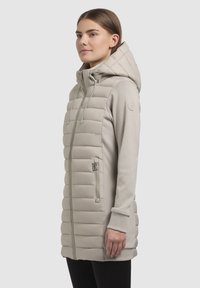 Lysbeige lang vest med en quiltet front, glatte ærmer, justerbar hætte og lynlåslomme på siden. Har ribbede manchetter.