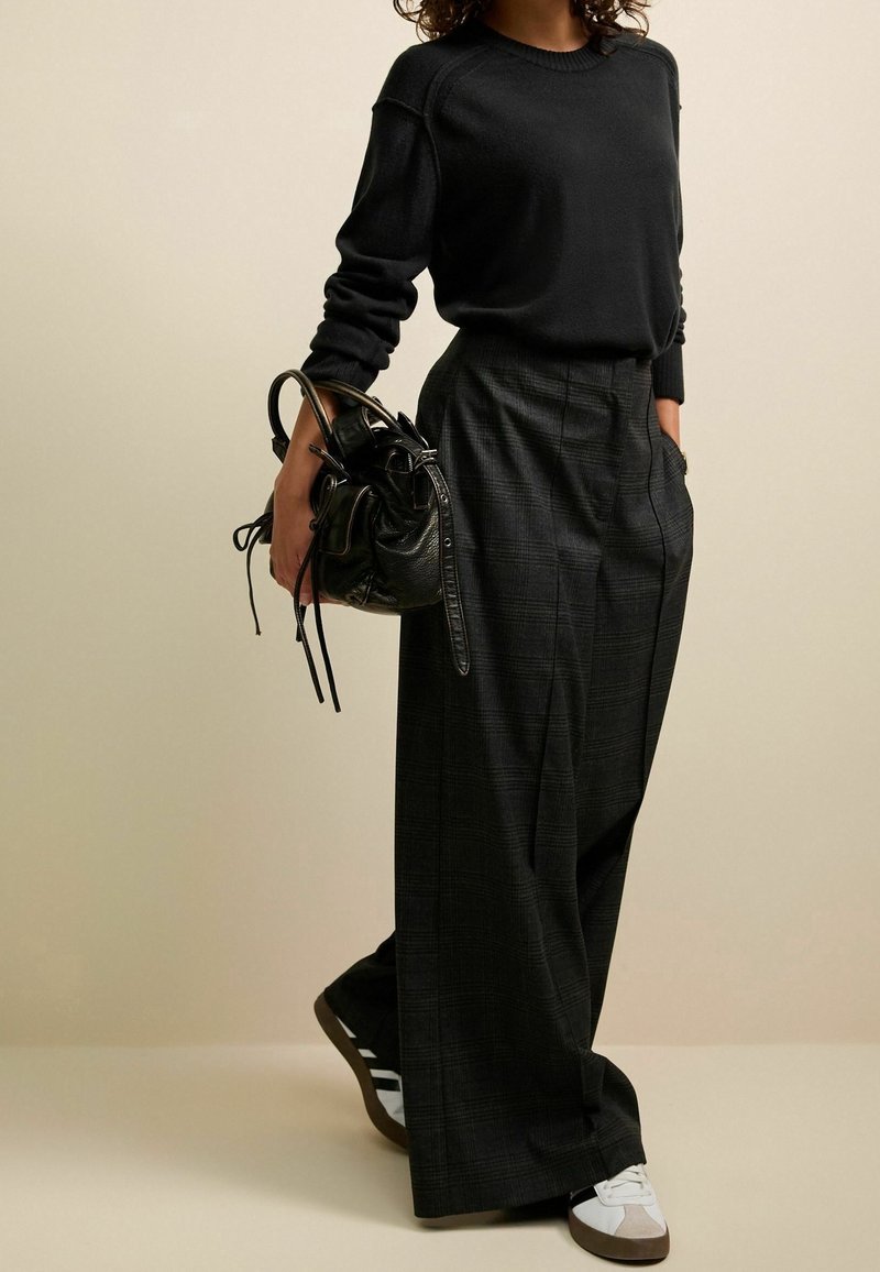 Personne portant un pull noir et un pantalon large à carreaux, tenant un sac à main en cuir noir et portant des baskets blanches sur un fond uni.
