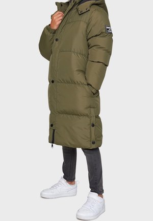 Person wearing an olive green long padded coat with hood, black skinny jeans, and white sneakers against a plain light background.