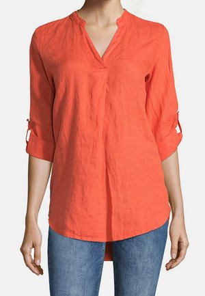 Frau trägt eine locker sitzende, langärmelige orange Bluse mit hochgekrempelten Ärmeln und blaue Jeans, steht vor einem einfarbigen Hintergrund.