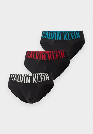Άνδρας μοντέλο με μαύρα εσώρουχα Calvin Klein στέκεται ξυπόλητος σε χαλί, ακουμπώντας το ένα χέρι σε μια μοντέρνα καρέκλα με μεταλλικά πόδια και μαξιλαρωμένο κάθισμα.