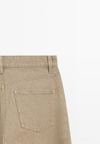 Beige Denimstoff, der eine Gesäßtasche mit sichtbaren Nähten und Gürtelschlaufen an einer Hose vor weißem Hintergrund zeigt.