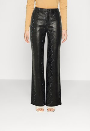 Pantalon noir taille haute à jambes larges avec un imprimé serpent discret, porté avec un haut beige à manches longues et des sandales blanches à talons ouverts.