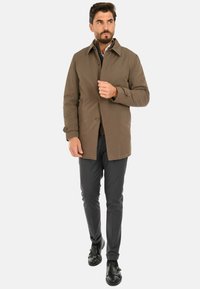 Giacca marrone leggera con collo a risvolto, chiusura con bottoni e maniche lunghe. Abbinata a pantaloni scuri e scarpe nere senza lacci.