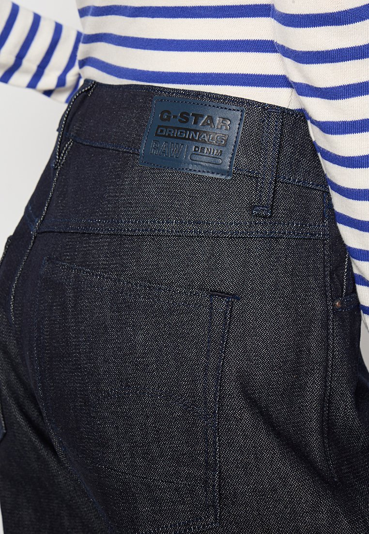 Jeans en denim foncé avec une surface texturée, arborant une étiquette en cuir bleu portant la mention "G-STAR ORIGINALS RAW DENIM" sur la ceinture.