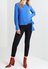 Kvinna i ljusblå långärmad blus med sidoknyte, svarta slim fit-byxor och rostfärgade klackstövletter som står framför en vit bakgrund.