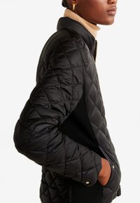 Veste matelassée noire avec un col montant, fermeture éclair avant et poches latérales, avec un tissu lisse contrastant avec un panneau texturé.