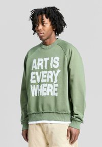 Felpa verde oliva con testo bianco logorato "L'ARTE È OVUNQUE", maniche raglan e orlo sfilacciato in fondo. Materiale in cotone.
