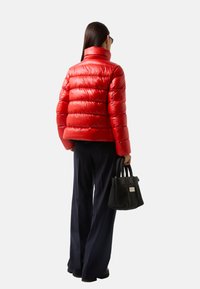 Doudoune rouge avec un col haut et un design matelassé, associée à un pantalon large bleu marine et un sac à main noir avec une étiquette.