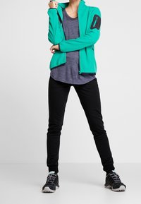 Heren zip-up jas van teal fleece met een zwart accentvak, gecombineerd met een grijs T-shirt en zwarte taps toelopende broek, en zwarte sportschoenen.