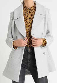 Vaaleanharmaa kaksirivinen takki, jossa on suuret kaulukset, valmistettu teksturoidusta kankaasta. Takki on kerrostettu ruskean leopardikuosisen paidan ja mustien farkkujen päälle.