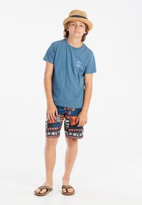 Camiseta de algodón azul, estampado gráfico en el pecho izquierdo; bañador con patrón colorido y varias formas. Sombrero de paja y chanclas marrones.