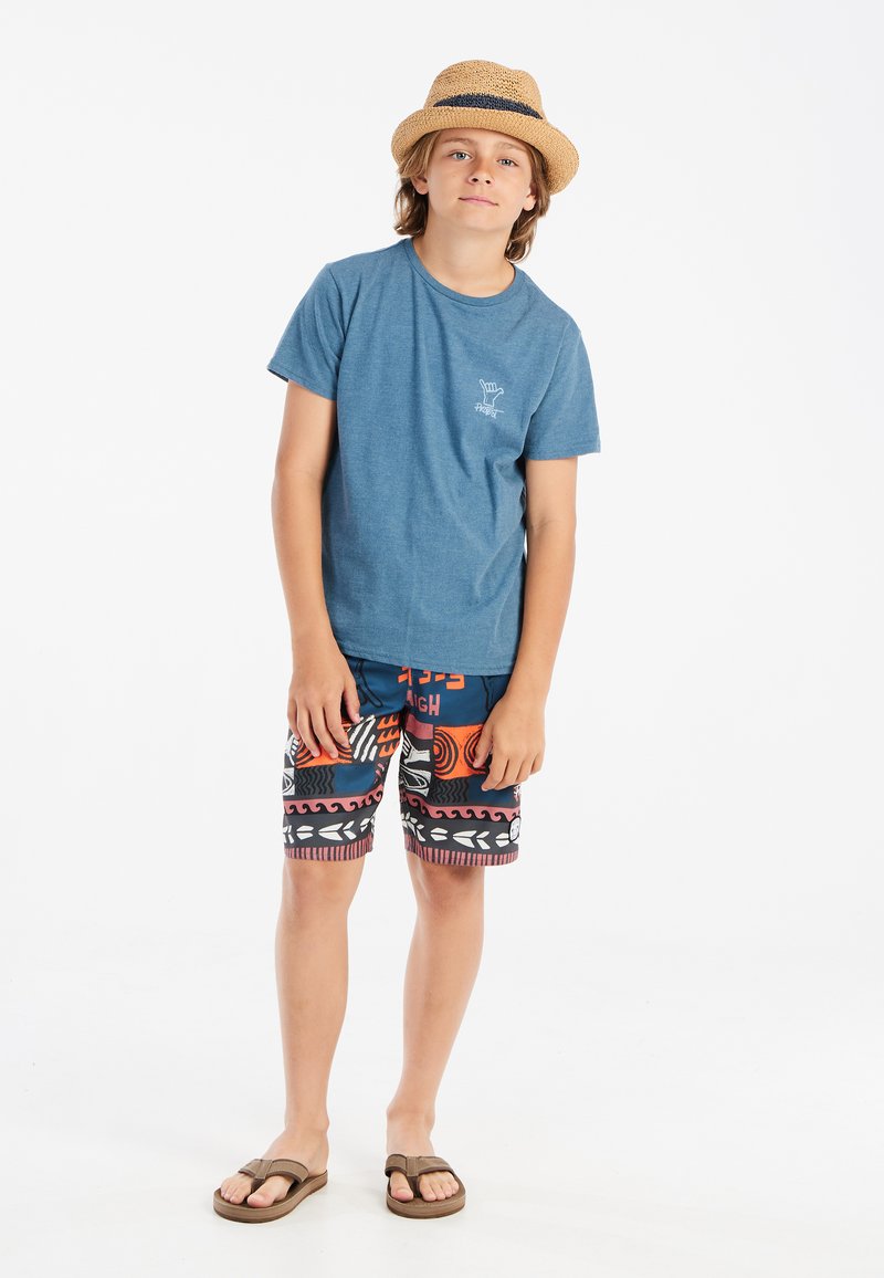 Camiseta de algodón azul, estampado gráfico en el pecho izquierdo; bañador con patrón colorido y varias formas. Sombrero de paja y chanclas marrones.